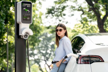 Yolculuk sırasında doğal orman ya da milli parktaki elektrikli araba için şarj olan genç bir kadın. Tatil ve tatil boyunca çevre dostu seyahat. Yükselt