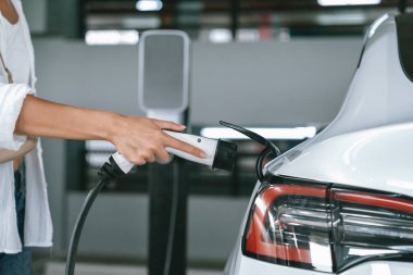 Genç bir kadın EV elektrikli arabasıyla alışveriş merkezinin otoparkına gidiyor şehir merkezinde elektrik iç organlarının yeşil temiz şarj edilebilir enerji ile sürdürülebilirlik yaşam tarzını gösteriyor.