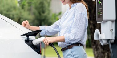 Açık yeşil şehir parkı manzaralı EV şarj istasyonundan genç kadın EV elektrikli araçları şarj ediyor. Çevre dostu şehir ulaşımı ve çevre dostu EV araba seyahati. Büyüleyici Panorama