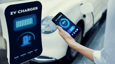 Akıllı telefonlu çalışan bir iş kadını akıllı EV mobil uygulama ile akü durumu arayüzünü tutarken EV arabası da otoparktaki şarj istasyonundan elektrik yüklüyor. İnceleyin