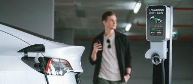 Genç adam EV elektrikli arabasıyla alışveriş merkezinin otoparkına gidiyor şehir merkezindeki şarj oluyor yeşil, temiz, yeniden şarj edilebilir elektrikli araç iç organları ile kentsel sürdürülebilirlik yaşam tarzını gösteriyor