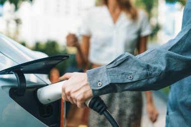 Genç çift yazın yeşil sürdürülebilir şehir bahçesinde EV elektrikli araba ile seyahat ederken, yeşil temiz yeniden şarj edilebilir elektrikli araç iç organları ile kentsel sürdürülebilirlik yaşam tarzını gösteriyor.
