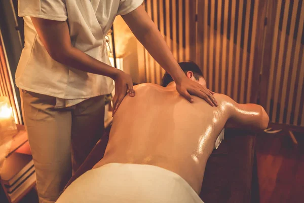 Rear view man customer enjoying relaxing anti-stress spa massage and pampering with beauty skin recreation leisure in warm candle lighting ambient salon spa at luxury resort or hotel. Quiescent