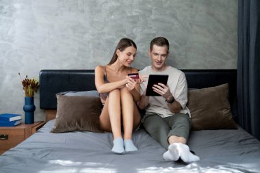 Genç çift, kredi kartıyla ödemek için internet üzerinden ödeme uygulaması ve dijital cüzdan kullanarak yatak odasında oturuyor. Mobil internet üzerinden alışveriş ve modern alımlar. Açılıyor
