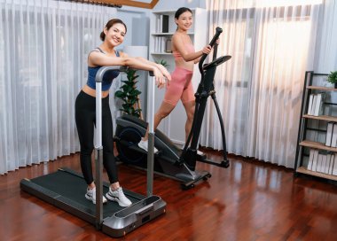 Enerjik ve güçlü atletik Asyalı kadın evde spor arkadaşı ya da antrenörü ile birlikte eliptik koşu makinesiyle koşuyor. Formda fiziğin takibi ve sağlıklı yaşam tarzına bağlılık. Kuvvetli