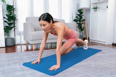 Spor kıyafetli Asyalı bir kadın antrenman rutini olarak spor minderinin üzerinde burpee yapıyor. Çekici kız sağlıklı yaşam tarzını ve formda vücut yapısını takip eder. Kuvvetli