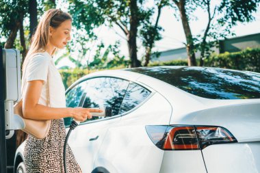 Genç bir kadın yazın yeşil sürdürülebilir şehir bahçesinde EV elektrikli arabasıyla seyahat ediyor. Kentsel sürdürülebilirlik yaşam tarzı yeşil temiz yeniden şarj edilebilir enerji elektrikli BEV araç iç organları