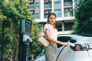 Genç bir kadın yazın yeşil sürdürülebilir şehir bahçesinde EV elektrikli arabasıyla seyahat ediyor. Kentsel sürdürülebilirlik yaşam tarzı yeşil temiz yeniden şarj edilebilir enerji elektrikli BEV araç iç organları