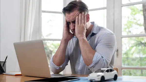 Stressed and frustrated car design engineer put his head in his hand after discover flaw in vehicle design, working at home for automotive business while trying to find solution with laptop.Synchronos