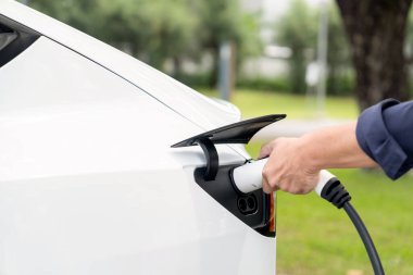Açık yeşil şehir parkı manzaralı EV şarj istasyonundan EV elektrikli araçları şarj eden adam. Çevre dostu şehir ulaşımı ve çevre dostu EV araba seyahati. Yükselt