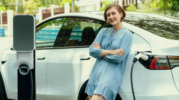 Beautiful young woman recharging her electric car from home EV charging station using alternative energy with net zero emission. Modern young girl charging her vehicle before vacation travel.Perpetual