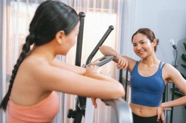 Enerjik ve güçlü atletik Asyalı kadın evde spor arkadaşı ya da antrenörü ile birlikte eliptik koşu makinesiyle koşuyor. Formda fiziğin takibi ve sağlıklı yaşam tarzına bağlılık. Kuvvetli