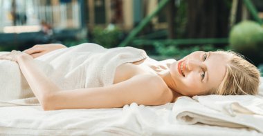 Güzel genç bir kadın, doğayla çevrili bir spa yatağında dinleniyor. Vücut masajına hazırım. Beyaz havluyla çekici bir kadın vücut masajı beklerken huzur içinde yatıyor. Kapatın. Huzur