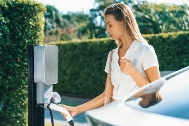 Genç bir kadın yazın yeşil sürdürülebilir şehir bahçesinde EV elektrikli arabasıyla seyahat ediyor. Kentsel sürdürülebilirlik yaşam tarzı yeşil temiz yeniden şarj edilebilir enerji elektrikli BEV araç iç organları