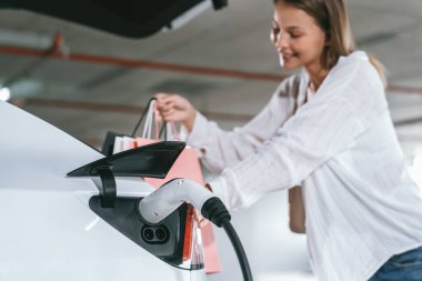 Genç bir kadın EV elektrikli arabasıyla alışveriş merkezinin otoparkına gidiyor şehir merkezinde elektrik iç organlarının yeşil temiz şarj edilebilir enerji ile sürdürülebilirlik yaşam tarzını gösteriyor.