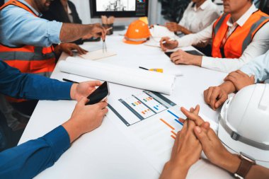 Mimari proje üzerinde birlikte çalışan çeşitli inşaat mühendisi ve müvekkil grupları, inşaat planını gözden geçirip toplantı masasında plan oluşturuyorlar. Tedbirli