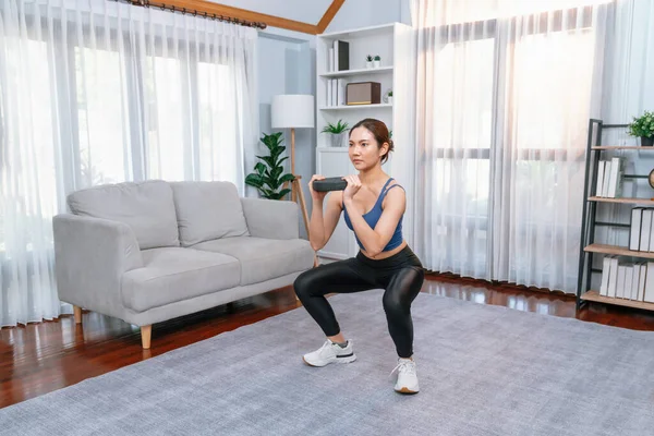 Vigorous energetic woman doing squat weight lifting exercise at home. Young athletic asian woman strength and endurance training session as home workout routine with squat.