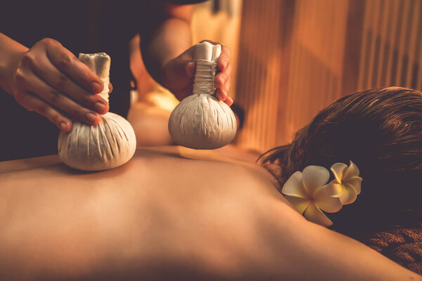 Hot herbal ball spa massage body treatment, masseur gently compresses herb bag on couple customer body. Serenity of aromatherapy recreation in warm lighting of candles at spa salon. Quiescent