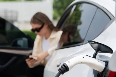 Genç bir kadın EV otomobil şarj istasyonu Green City Park 'ta elektrik ödemek için akıllı telefonu kullanıyor. Modern çevre ve EV araçlı sürdürülebilir şehir yaşam tarzı. Yararlı