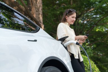 Genç bir kadın EV otomobil şarj istasyonu Green City Park 'ta elektrik ödemek için akıllı telefonu kullanıyor. Modern çevre ve EV araçlı sürdürülebilir şehir yaşam tarzı. Yararlı