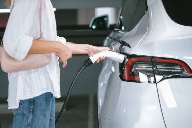Genç bir kadın EV elektrikli arabasıyla alışveriş merkezinin otoparkına gidiyor şehir merkezinde elektrik iç organlarının yeşil temiz şarj edilebilir enerji ile sürdürülebilirlik yaşam tarzını gösteriyor.