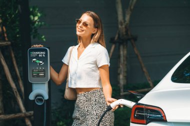 Genç bir kadın yazın yeşil sürdürülebilir şehir bahçesinde EV elektrikli arabasıyla seyahat ediyor. Kentsel sürdürülebilirlik yaşam tarzı yeşil temiz yeniden şarj edilebilir enerji elektrikli BEV araç iç organları