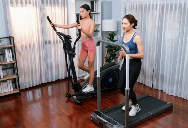 Enerjik ve güçlü atletik Asyalı kadın evde spor arkadaşı ya da antrenörü ile birlikte eliptik koşu makinesiyle koşuyor. Formda fiziğin takibi ve sağlıklı yaşam tarzına bağlılık. Kuvvetli