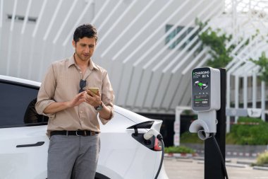 Genç adam, alışveriş merkezinin otoparkındaki EV otomobil şarj istasyonunda elektriği ödemek için akıllı telefonu kullanıyor. Modern çevre ve EV araçlı sürdürülebilir şehir yaşam tarzı. Yararlı