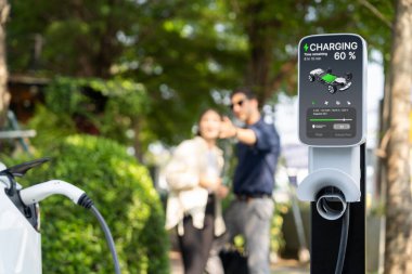Genç çift, baharda açık yeşil şehir parkındaki şarj istasyonundan elektrikli araba aküsünü şarj ediyor. Sürdürülebilir çevre dostu şehir seyahati için şarj edilebilir EV araba.