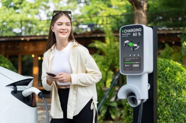 İlkbahar bahçesindeki açık kafede çalışan EV elektrikli araba şarjı, yeşil şehir sürdürülebilirliği ve çevre dostu EV otomobili için genç bir kadınla sürdürülebilir şehir seyahati elektrik ödüyor. Yararlı