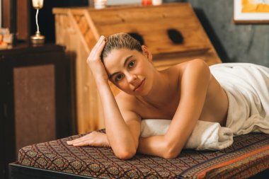 Güzel genç bir kadın, sauna dolabının önündeki spa yatağında yatıyor. Beyaz havluyla beyaz havluyla, sauna odasında vücut masajı beklerken kameraya bakan çekici, rahatlatıcı beyaz kadınlar. Huzur..