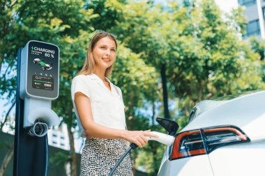 Genç bir kadın yazın yeşil sürdürülebilir şehir bahçesinde EV elektrikli arabasıyla seyahat ediyor. Kentsel sürdürülebilirlik yaşam tarzı yeşil temiz yeniden şarj edilebilir enerji elektrikli BEV araç iç organları