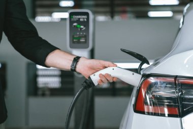 Genç adam EV elektrikli arabasıyla alışveriş merkezinin otoparkına gidiyor şehir merkezindeki şarj oluyor yeşil, temiz, yeniden şarj edilebilir elektrikli araç iç organları ile kentsel sürdürülebilirlik yaşam tarzını gösteriyor
