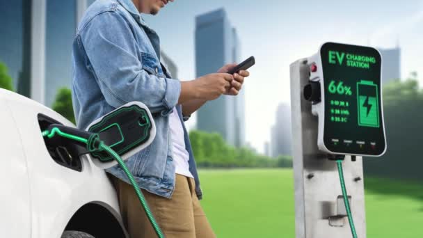 亚洲人在绿色城市公园等着用智能手机充电 而他的电动汽车则从未来的充电站充电 智能和未来主义绿色能源基础设施 仔细阅读 — 图库视频影像