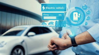 İşadamı, modern bina arka planına sahip şarj istasyonundan şarj olurken akıllı saat hologramının EV akü durumunu kontrol ediyor. EV teknolojisi adaptasyonu için temiz enerjinin gelecekteki yaşam tarzı. İnceleyin