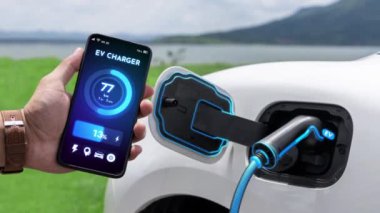 Akıllı telefon ekran akü durumu arayüzünü elinde tutan iş adamı akıllı EV mobil uygulama ile EV otomobili elektrik şarj istasyonundan doğal göl arka planı ile elektrik. İnceleyin.