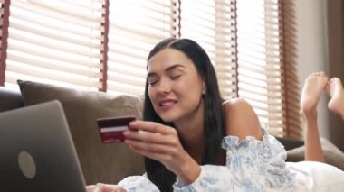 Kanepede yatan genç bir kadın internet bankacılığı için kredi kartıyla dizüstü bilgisayar kullanıyor. Evdeki online ödeme kapısından alışveriş yapıyor. Modern ve uygun online satın alma. Blithe