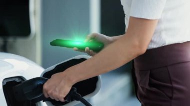 Modern yaşam tarzı bir kadın elektrikli arabasına EV şarj aleti yerleştiriyor ve akıllı telefondan EV batarya durum hologramını kontrol ediyor. Gelecekçi enerji sürdürülebilirliği ve elektrikli araba. İnceleyin