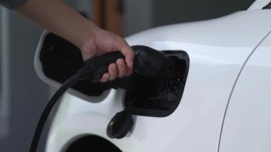 Adam elektrikli arabasını akıllı ev şarj istasyonundaki EV şarj cihazıyla şarj ediyor. Fütürist dijital batarya durumu hologramı. Sürdürülebilir güç için EV araba ve ev enerjisi altyapısı. İnceleyin