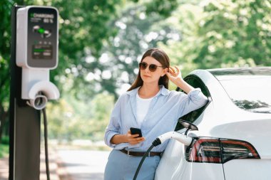Ulusal Park veya Yaz Ormanı 'ndaki tatil gezisi sırasında EV şarj istasyonundan elektrik aküsü almak için akıllı telefon internet bankacılık uygulaması kullanan genç bir kadın. Yükselt
