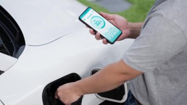 Modern yaşam tarzı adamı EV şarj cihazını ev şarj istasyonuna yerleştirir ve akıllı telefondan EV batarya durumunu etkinleştirir. Gelecekçi enerji sürdürülebilirliği ve elektrikli araba. İnceleyin