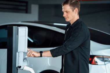 Genç adam EV elektrikli arabasıyla alışveriş merkezinin otoparkına gidiyor şehir merkezindeki şarj oluyor yeşil, temiz, yeniden şarj edilebilir elektrikli araç iç organları ile kentsel sürdürülebilirlik yaşam tarzını gösteriyor