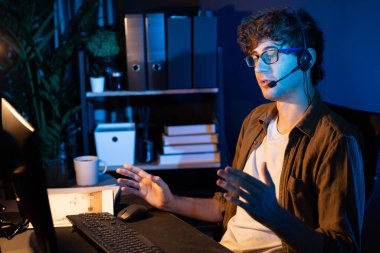 Genç yaratıcı iş adamı, bilgisayar ekranında dinleyicilerle iletişim kuruyor ya da takım üyesiyle görüşüyor. Neon Light Modern Ev Ofisi 'nde kulaklık takıyor. Gece vakti çalışma kavramı. Toplayıcı.