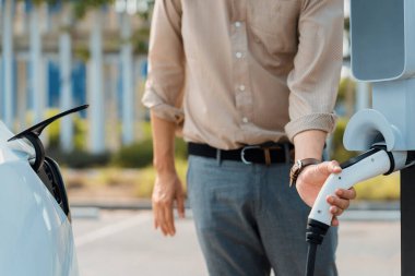 Genç adam, şehrin ticari otoparkındaki şarj istasyonundan elektrikli araba aküsünü şarj etmek için EV şarj aleti koymuş. Sürdürülebilir çevre dostu şehir yolculuğu için şarj edilebilir EV araba. Yararlı