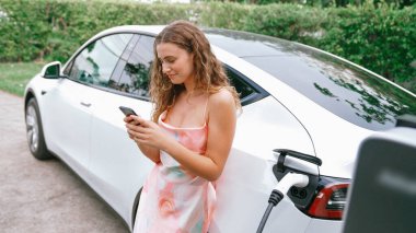 Genç bir kadın doğadaki EV şarj istasyonunda elektrik için akıllı telefon kullanıyor. Modern çevre ve EV araçlı sürdürülebilir otomobil taşıma yaşam tarzı. Eşzamanlama