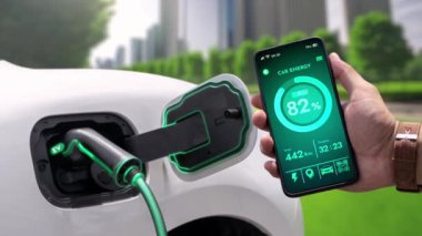 İşadamı çevre yeşil şehir parkındaki şarj istasyonundan şarj olurken akıllı telefon ekranından EV batarya durum mobil uygulamasını kontrol ediyor. Elektrikli arabanın gelecekteki enerji sürdürülebilirliği. İnceleyin