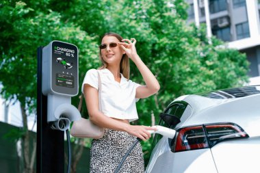 Genç bir kadın yazın yeşil sürdürülebilir şehir bahçesinde EV elektrikli arabasıyla seyahat ediyor. Kentsel sürdürülebilirlik yaşam tarzı yeşil temiz yeniden şarj edilebilir enerji elektrikli BEV araç iç organları
