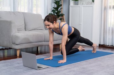 Spor kıyafetli Asyalı bir kadın antrenman rutini olarak spor minderinin üzerinde burpee yapıyor. Çekici kız sağlıklı yaşam tarzını online egzersiz videosuyla takip ediyor. Kuvvetli