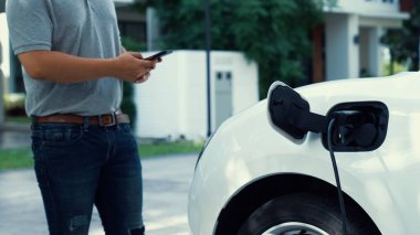 Elektrikli araba şarj istasyonuna bağlanıyor. Arka planda akıllı telefon kullanan adam EV şarj cihazıyla şarj oluyor. Sürdürülebilir temiz enerji kullanımının gelecekteki yaşam tarzı. İnceleyin