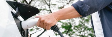 Yolculuk sırasında doğal orman ya da milli parktaki elektrikli araba için şarj olan bir adam. Tatil ve tatil boyunca çevre dostu seyahat. Panorama Sancağını Yükselt